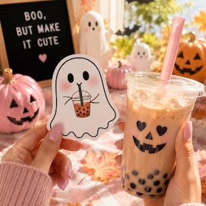 May include: A die-cut sticker featuring a white ghost with pink cheeks holding a bubble tea, alongside a clear plastic cup filled with milk tea and dark boba pearls, decorated with a black heart-eyed jack-o'-lantern face and a pink straw.