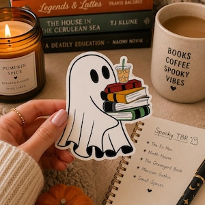 May include: A die-cut vinyl sticker featuring a white ghost with black eyes holding a stack of four colorful books and a plastic cup with a straw. The ghost has a simple outline and is depicted in a cartoon style with a light-colored background.