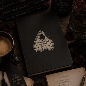 May include: A black notebook featuring a white planchette-shaped sticker on the cover. The sticker displays the text "summoning my last fuck" surrounded by small star and cross symbols. The notebook has a smooth, matte black cover and a rectangular shape.