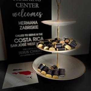 May include: A tiered serving stand with gold detailing displays wrapped chocolates. A sign in the background reads "The Home Missionary Training Center welcomes Hermana Zabriskie." A card with a map of Costa Rica is also visible.