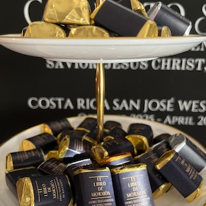 Può includere: Un vassoio a due livelli contiene cioccolatini confezionati. Il ripiano superiore presenta cioccolatini avvolti in oro, mentre il ripiano inferiore mostra cioccolatini avvolti in blu scuro e oro con testo in spagnolo. Il vassoio ha uno stelo dorato e un bordo bianco e oro.