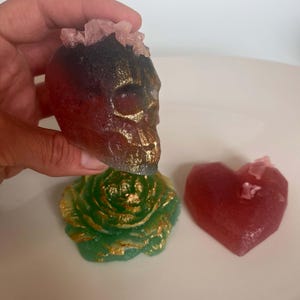 Könnte beinhalten: Sammlung dekorativer Resin-Objekte: ein Schädel, eine Rose und ein Herz. Der Schädel ist rot und goldfarben mit einer rosa Kristallkrone. Die Rose ist grün mit goldenen Akzenten. Das Herz ist rot mit einem rosa Kristall.