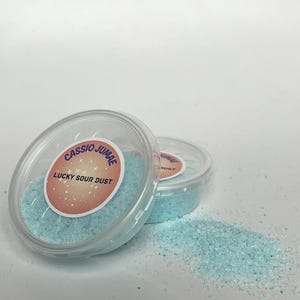 Könnte beinhalten: Zwei transparente Plastikbehälter gefüllt mit hellblauem Pulver, beschriftet mit "Lucky Sour Dust" und dem Markennamen "Cassio Jomae". Ein kleiner Haufen des gleichen Pulvers ist in der Nähe verstreut. Der Hintergrund ist schlicht weiß.