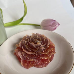 Pode incluir: Um objeto decorativo em forma de rosa, com tons de vermelho e roxo, acentuado com glitter dourado. O design floral é apresentado em um prato branco com borda dourada, com uma tulipa e vegetação ao fundo.