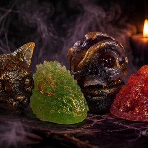 Op de afbeelding: Vier decoratieve objecten waaronder een bronskleurige kattenkop, een doorschijnend groen getextureerd wezen, een donkere schedel omwikkeld met goudkleurige spiralen en een piramidevormige cluster van kleine rode bollen in een donkere setting.