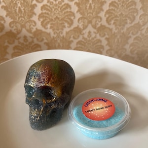 Peut inclure: Une figurine de crâne pailletée et multicolore aux tons sombres de noir, d'or et de rouge, placée à côté d'un petit récipient en plastique transparent rempli de poudre bleu clair, avec une étiquette indiquant CASSIO JUMAE LUCKY SOUR DUST.