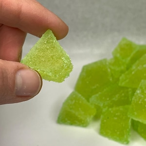 Può includere: Una mano tiene in mano una singola caramella gommosa triangolare verde lime spolverata di zucchero. Diverse caramelle simili sono sullo sfondo, suggerendo un lotto più grande. Le caramelle hanno un aspetto leggermente traslucido.