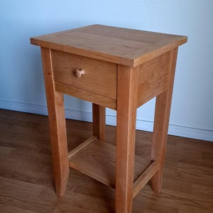 May include: A small, wooden side table with a single drawer and a lower shelf. The table is made of light-colored wood and has a simple, classic design. The table measures approximately 24 inches tall, 18 inches wide, and 18 inches deep.