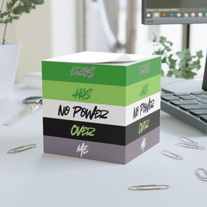 Puede incluir: Un bloc de notas cúbico con el texto "EROS HAS NO POWER OVER ME" en verde, negro, blanco y gris. El bloc de notas está sobre una superficie blanca con clips esparcidos, un portátil y unas tijeras.