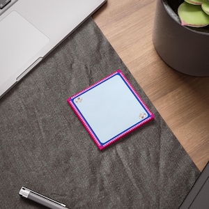 May include: A square notepad with a light blue interior, a dark blue border, and a pink decorative edge. The notepad has small illustrations in each corner. The notepad is on a dark gray surface, near a laptop and a pen.
