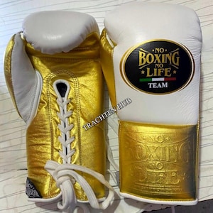 May include: Pair of boxing gloves with a gold and white color scheme. The gloves feature a lace-up design and the text "NO BOXING NO LIFE TEAM" on the wrist. The gloves are designed for boxing.