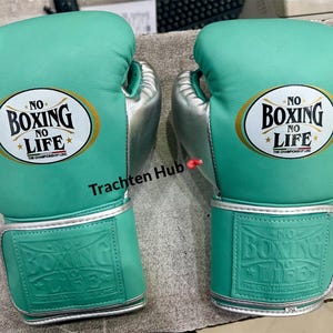 May include: A pair of teal boxing gloves with silver accents. Each glove features the text "NO BOXING NO LIFE" in a gold oval. The gloves are designed for boxing and combat sports.
