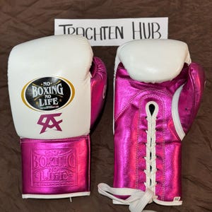 Puede incluir: Par de guantes de boxeo rosas y blancos. Los guantes tienen el texto "NO BOXING NO LIFE" y un logotipo en la parte delantera. Los guantes están atados con cordones blancos y tienen una correa de muñeca rosa.