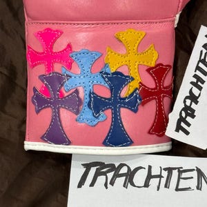 Op de afbeelding: Roze leren bokshandschoen met een wit kruisontwerp en kleurrijke kruispatches op de polsband.