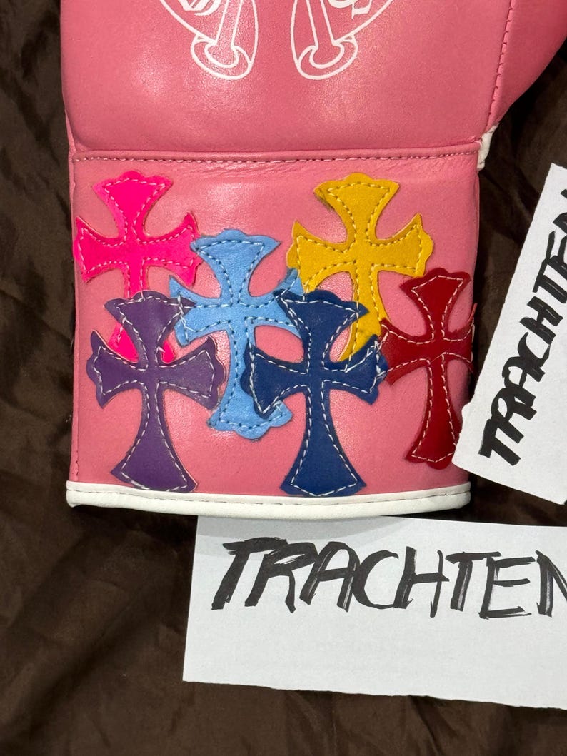 Op de afbeelding: Roze leren bokshandschoen met een wit kruisontwerp en kleurrijke kruispatches op de polsband.