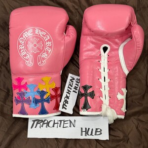 Op de afbeelding: Een paar roze bokshandschoenen met witte veters. E&eacute;n handschoen heeft een wit Chrome Hearts logo en de andere heeft kleurrijke kruisontwerpen.
