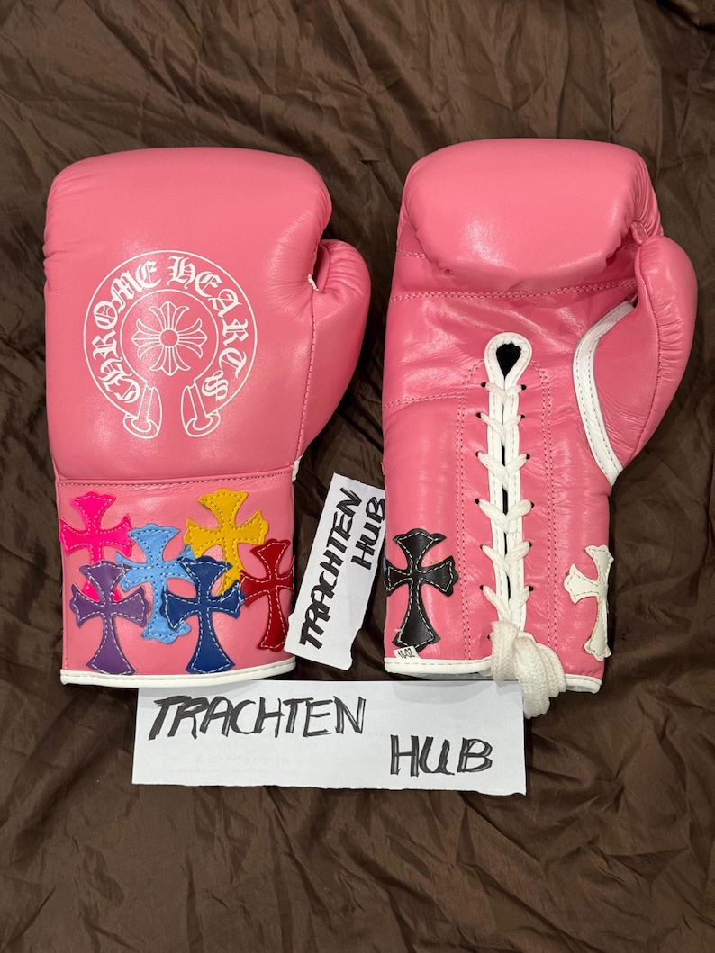 Op de afbeelding: Een paar roze bokshandschoenen met witte veters. E&eacute;n handschoen heeft een wit Chrome Hearts logo en de andere heeft kleurrijke kruisontwerpen.