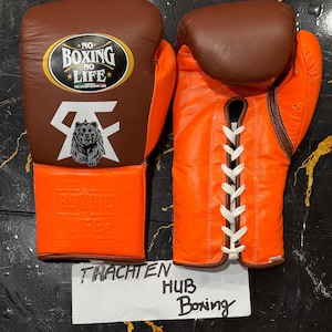 May include: A pair of boxing gloves in brown and orange leather. The left glove features a logo with the text "NO BOXING NO LIFE" and a bear graphic. The right glove has white laces. The gloves are on a dark surface.