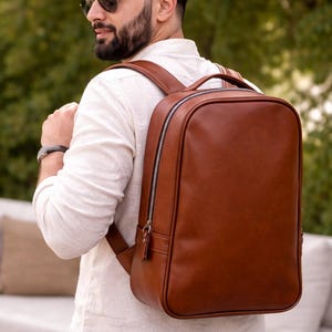 Könnte beinhalten: Ein brauner Lederrucksack mit rechteckiger Form und schwarzem Reißverschluss. Der Rucksack hat einen oberen Griff und verstellbare Schultergurte. Die Person im Bild trägt ein weißes Hemd und eine Sonnenbrille.