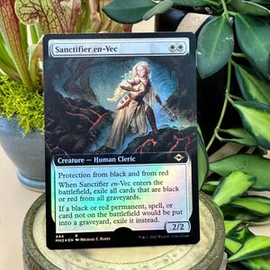 May include: A Magic: The Gathering card featuring "Sanctifier en-Vec." The card depicts a human cleric with a sword in a fantasy setting. The card has a silver border and includes game text about protection from black and red.