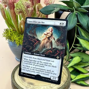 May include: A Magic: The Gathering card titled "Sanctifier en-Vec" featuring a Human Cleric illustration. The card has a white border with black text describing its abilities and stats. The background includes a fantasy landscape with red and black elements.