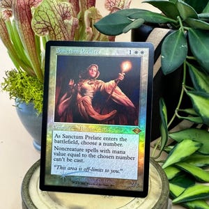 May include: A Magic: The Gathering card titled "Sanctum Prelate." The card features a woman in a flowing robe holding a torch. The card has a holographic sheen and includes game text. The background includes plants.