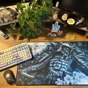 May include: A desk setup featuring a keyboard, mouse, and a large mousepad with a samurai warrior design in shades of blue and white. The desk also includes decorative items, plants, and a small painting of a cat.