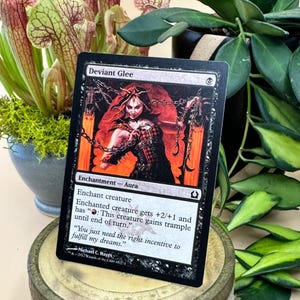 May include: A black trading card titled "Deviant Glee" featuring a red and black illustration of a woman with chains. The card is an enchantment-aura with text describing its effects. The card is displayed in front of green plants.