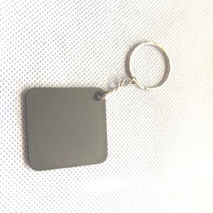 May include: A grey square keyring with a silver metal ring. The keyring is made of a textured material.