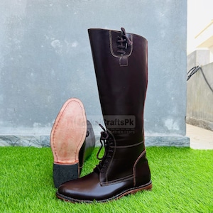 Puede incluir: Botas de montar de cuero marrón oscuro con cordones y caña alta. Las botas se exhiben sobre una superficie verde, con una bota en posición vertical y otra que muestra la suela. El texto "CraftsPk Leather Item's" es visible en el lateral de la bota.