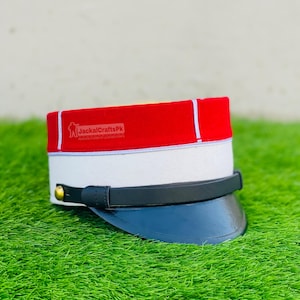 Könnte beinhalten: Ein roter, weißer und schwarzer Hut mit schwarzem Visier. Der Hut hat ein weißes Band und eine rote Oberseite mit weißer Zierleiste. Der Text "JackalCraftsPk" ist auf dem Hut zu sehen. Der Hut liegt auf grünem Gras.