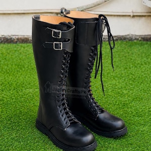 Puede incluir: Un par de botas negras de cuero hasta la rodilla. Las botas tienen un diseño con cordones, dos correas con hebilla y una suela robusta. Las botas se muestran sobre una superficie verde.