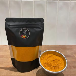 Puede incluir: Una bolsa negra resellable con una etiqueta plateada y el texto "ATRIYA" en blanco. La bolsa contiene un polvo amarillo brillante. Un pequeño cuenco de metal lleno del mismo polvo amarillo se encuentra sobre una superficie de madera.