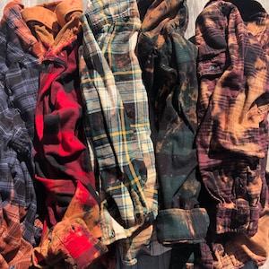 May include: Six plaid flannel shirts in various colors and patterns, including blue, red, green, and brown. The shirts are folded and arranged in a row on a wooden surface.