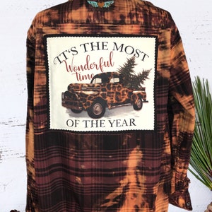 May include: A brown and orange plaid flannel shirt with a patch on the back that says "It's the most wonderful time of the year" with a vintage truck carrying a Christmas tree.