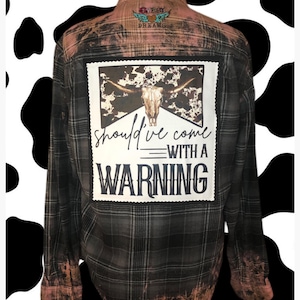 May include: Black and white plaid flannel shirt with a bleached effect. The back of the shirt features a patch with a cow skull and the text "should've come with a warning".