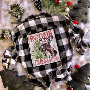 May include: Black and white plaid flannel shirt with a Christmas-themed graphic on the back. The graphic features a cowboy riding a bucking bronco near a Christmas tree, with the text "Rockin' Around the Christmas Tree".