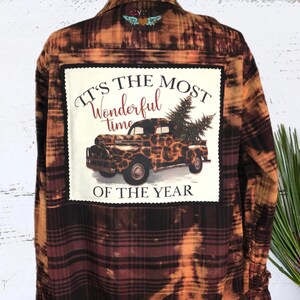 May include: A brown and orange plaid flannel shirt with a patch on the back that says "It's the most wonderful time of the year". The patch features a vintage truck with a Christmas tree in the back and a leopard print design.