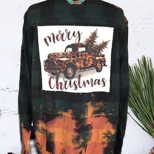 May include: A green and orange plaid flannel shirt with a white patch on the back that says "Merry Christmas" and features a vintage truck with a Christmas tree in the bed. The truck is decorated with a leopard print pattern.