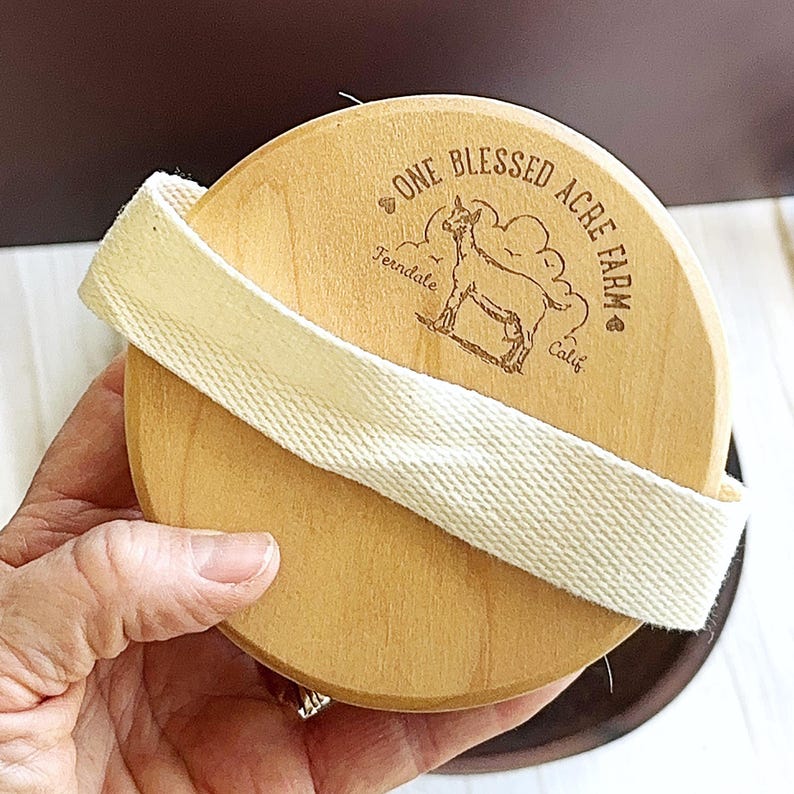 Puede incluir: Un cepillo corporal redondo de madera clara con una correa de tela color crema en el centro. La superficie de madera est&aacute; grabada con l&aacute;ser con una ilustraci&oacute;n de una cabra y el texto One Blessed Acre Farm, Ferndale, Calif.