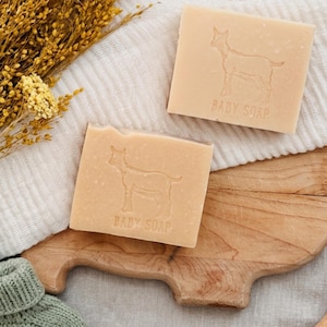 Puede incluir: Dos pastillas de jabón para bebé cuadradas con una silueta de cabra y la inscripción "BABY SOAP". El jabón es de color beige claro y está sobre una tabla de cortar de madera y un paño blanco, con flores amarillas secas en el fondo.
