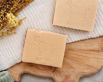 Puede incluir: Dos pastillas de jabón para bebé cuadradas con una silueta de cabra y la inscripción "BABY SOAP". El jabón es de color beige claro y está sobre una tabla de cortar de madera y un paño blanco, con flores amarillas secas en el fondo.