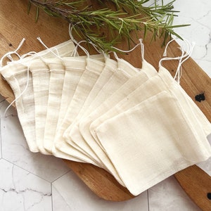 May include: A collection of beige muslin tea bags with drawstring closures, arranged on a wooden cutting board. Fresh rosemary sprigs are placed on the board. The tea bags are neatly fanned out, showcasing their simple, natural design.