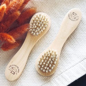 May include: Two wooden brushes with light-colored handles and natural bristles. Each handle features a stamped logo with the text One Blessed Acre Farm and a goat illustration. The brushes are positioned side by side on a textured fabric surface.