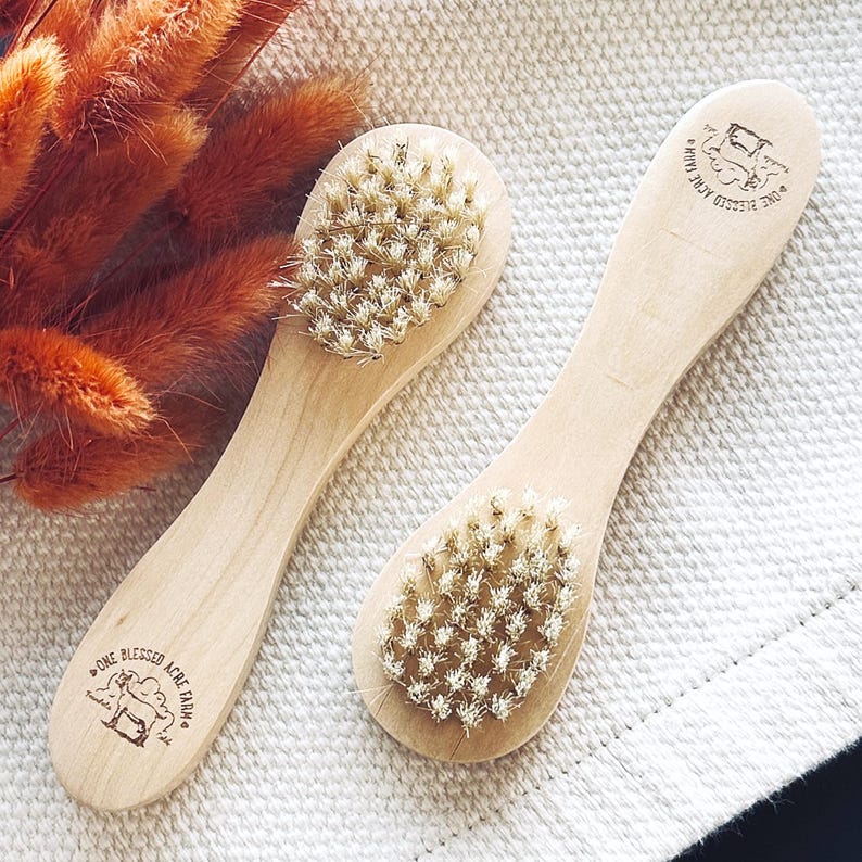 May include: Two wooden brushes with light-colored handles and natural bristles. Each handle features a stamped logo with the text One Blessed Acre Farm and a goat illustration. The brushes are positioned side by side on a textured fabric surface.