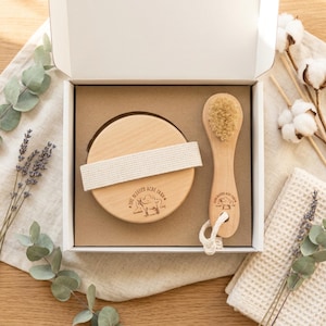May include: A white gift box containing a round wooden body brush with a beige fabric strap and a smaller wooden facial brush with natural bristles. Both brushes feature a stamped logo of a goat and the text One Blessed Acre Farm.