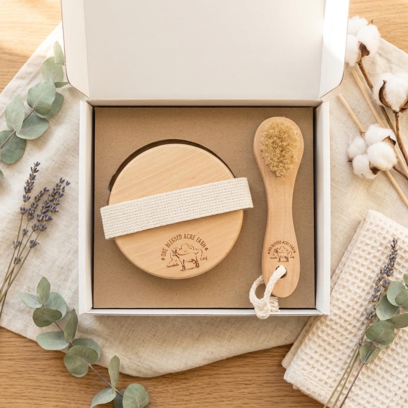 May include: A white gift box containing a round wooden body brush with a beige fabric strap and a smaller wooden facial brush with natural bristles. Both brushes feature a stamped logo of a goat and the text One Blessed Acre Farm.
