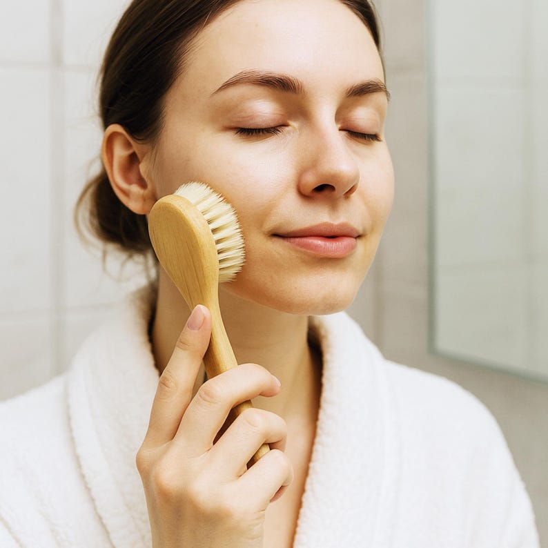 Puede incluir: Un cepillo de limpieza facial con mango de madera de color claro y cerdas suaves, densas y de color blanco roto. El cepillo presenta un cabezal de forma ovalada y un mango liso y ergon&oacute;mico dise&ntilde;ado para uso manual sobre la piel del rostro.