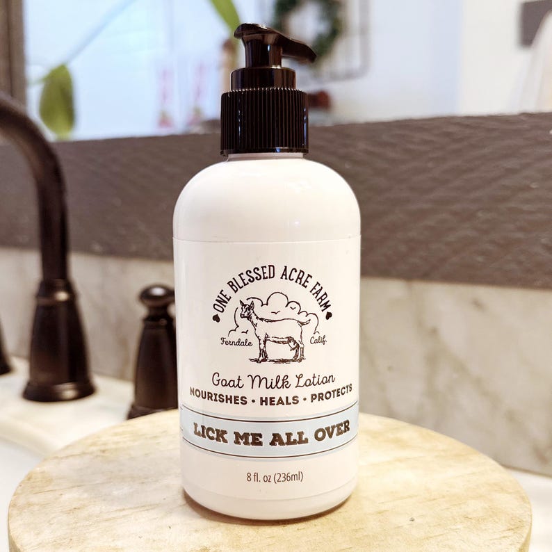 K&ouml;nnte beinhalten: Wei&szlig;e Flasche Ziegenmilch-Lotion mit schwarzem Pumpspender. Das Etikett tr&auml;gt die Aufschrift "One Blessed Acre Farm" mit einer Ziegenillustration und dem Text "LICK ME ALL OVER". Die Flasche enth&auml;lt 236 ml Lotion.