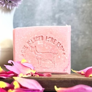 May include: A rectangular bar of pink soap featuring an embossed design of a goat and the text "ONE BLESSED ACRE FARM" centered on the front surface. The soap has a smooth, solid texture and is surrounded by scattered pink and yellow flower petals.